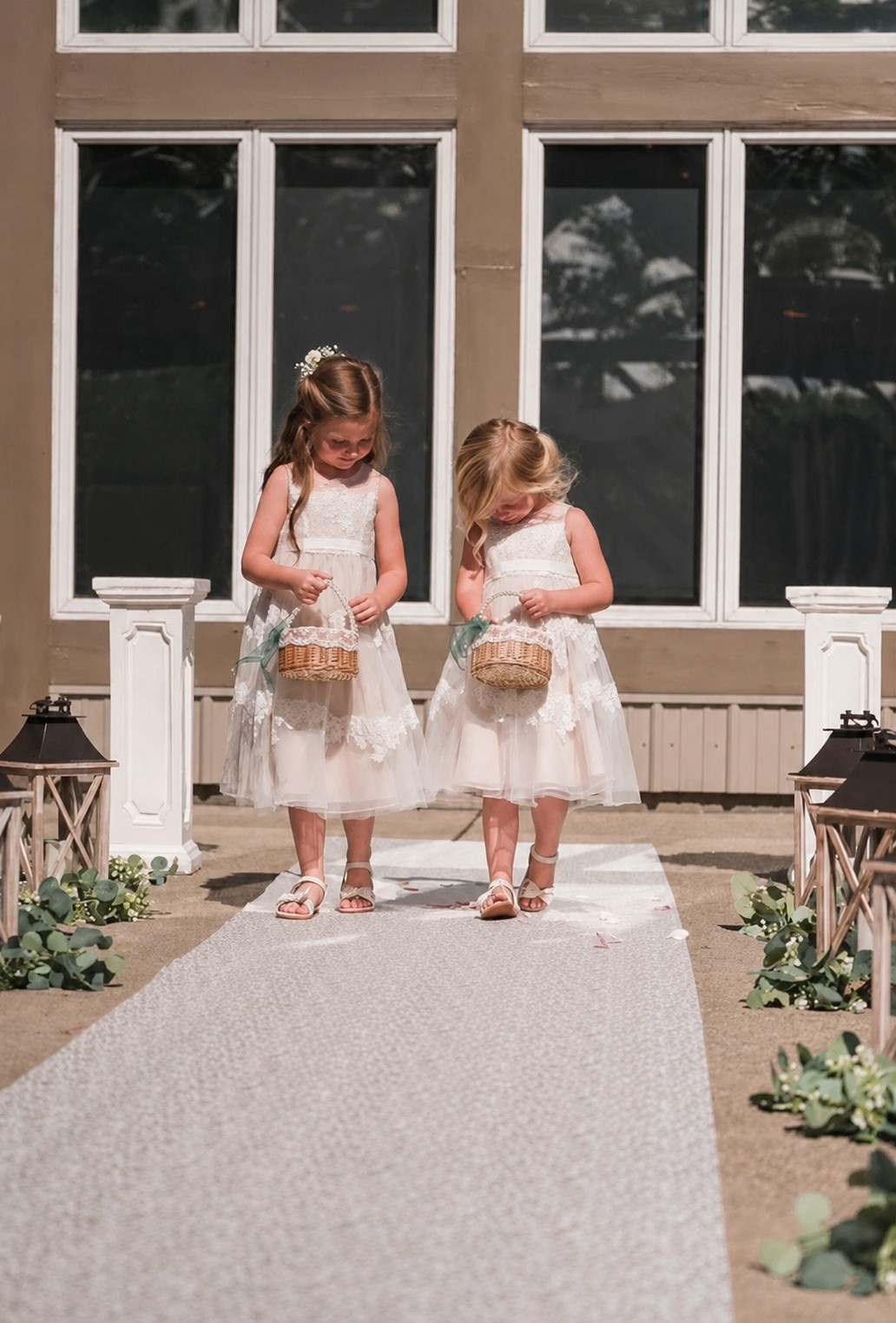 Two flower girls walking along a wedding ceremony aisle lined with lanterns and greenery