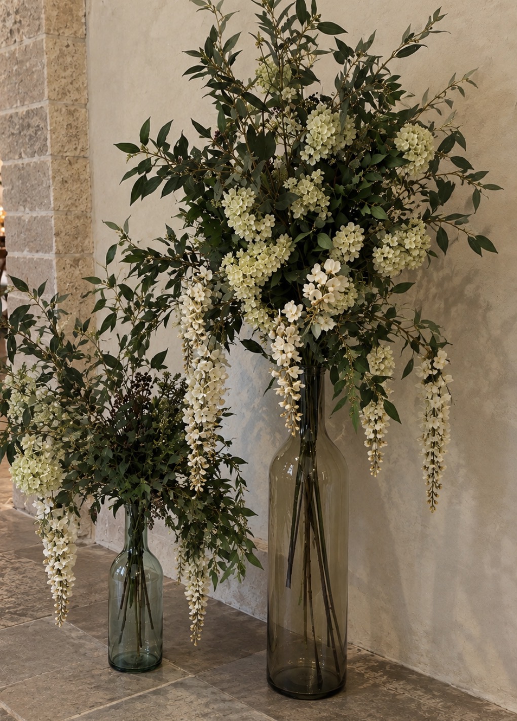 Tall glass floor vases filled with airy white flowers and abundant greenery